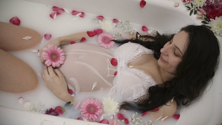 A serene pregnant woman relaxes in a milk bath adorned with pink flowers, symbolizing maternity. 15 Self-Care Ideas for the Exhausted Pregnant Woman