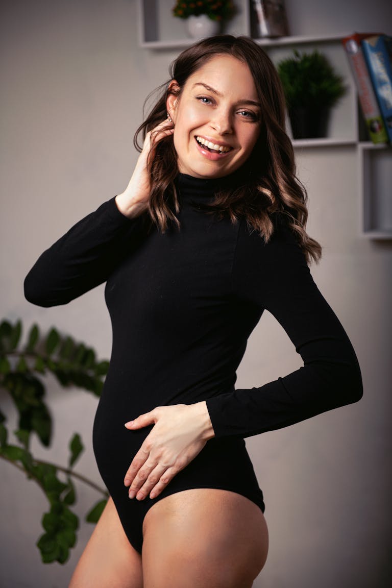 Happy pregnant woman in a black bodysuit poses indoors, touching her belly and smiling.