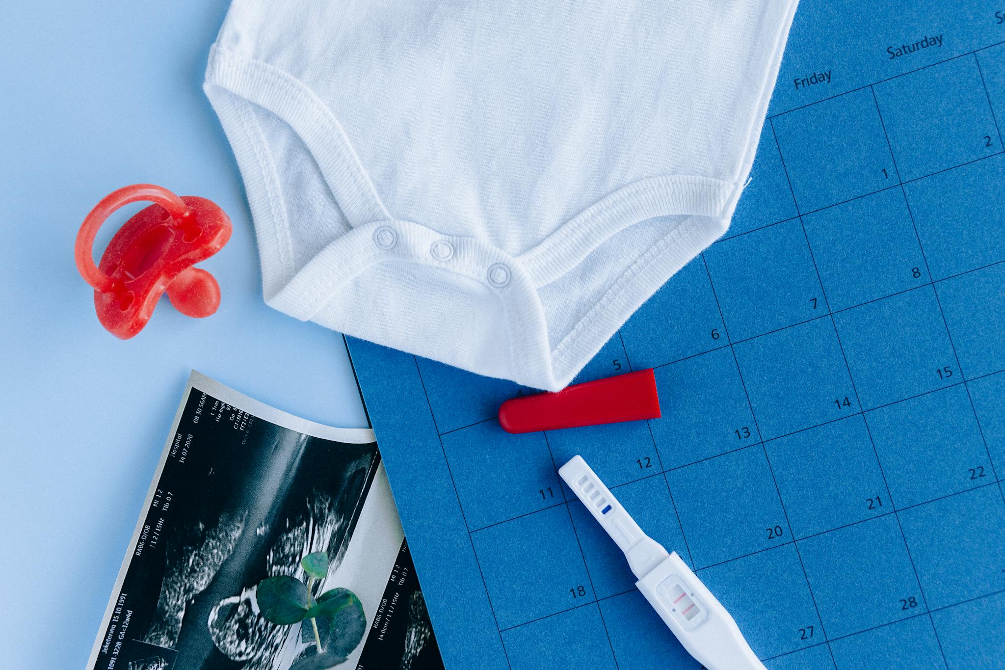 Pregnancy test, baby clothes, and calendar on blue surface symbolize planning for a baby's arrival.