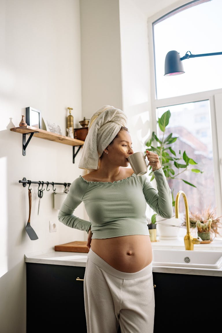 Pregnant woman in kitchen enjoying a hot drink, embracing relaxation and comfort. 🥑 Pregnancy Superfoods You Didn’t Know About