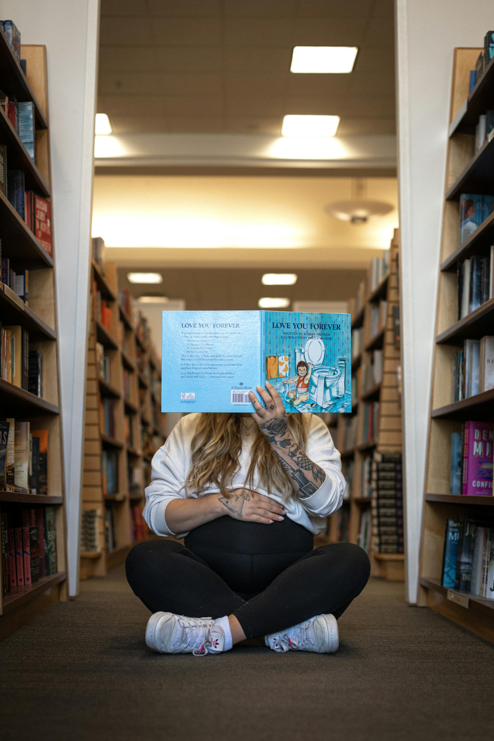 Pregnant woman sitting cross-legged reading a storybook in a library aisle.