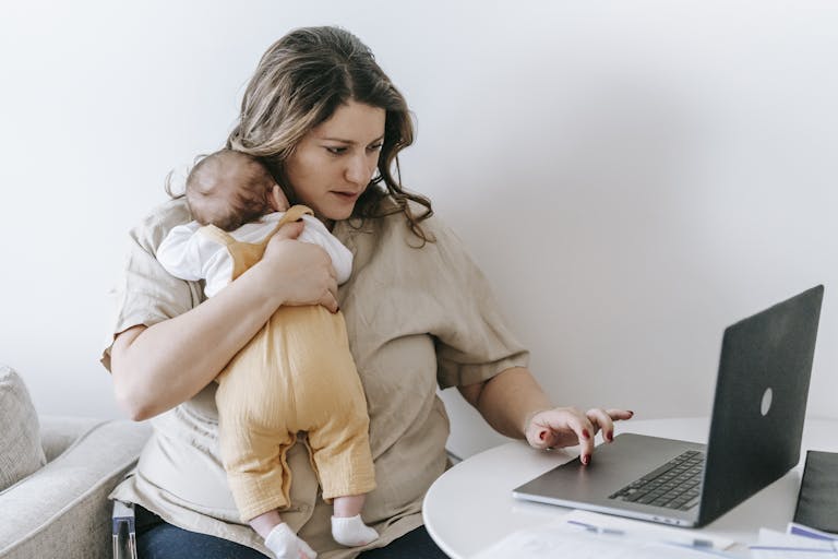 Concentrated young female freelancer embracing newborn while sitting at table and working remotely on laptop at home The Silent Struggles of Modern Motherhood: What No One Talks About