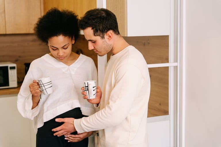 Interracial couple shares a tender moment with coffee mugs, embracing a pregnant belly.