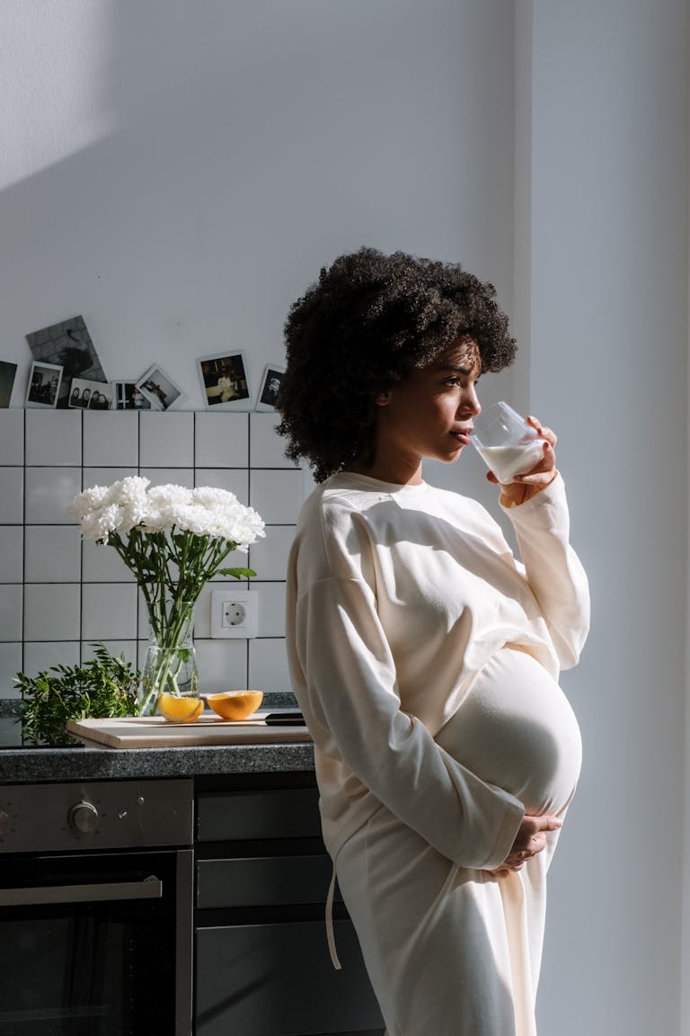 Pregnant woman drinking milk in a sunlit kitchen, surrounded by flowers and fruit.
