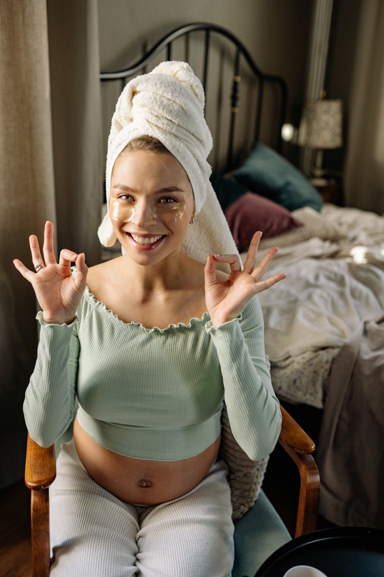 Pregnant woman smiling with skincare patches and towel on head in cozy bedroom. https://momtobe.store/pregnancy-skincare-routine-safe-products/