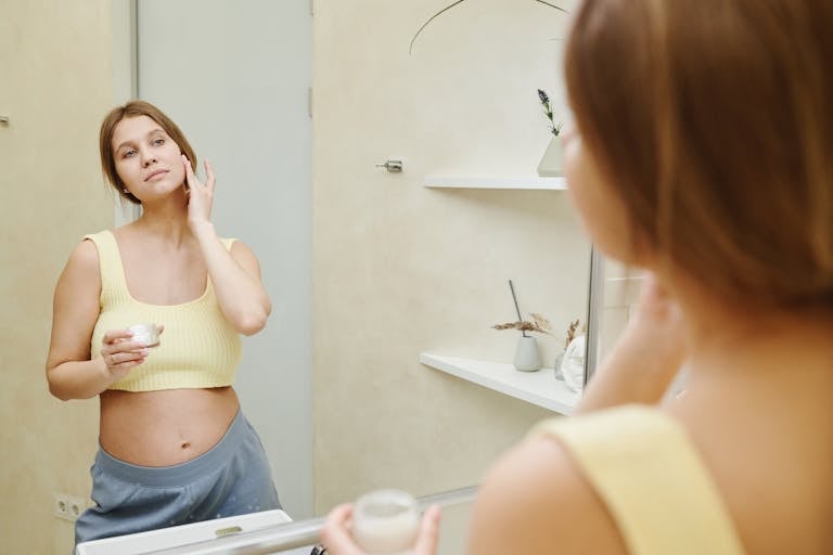 Pregnant woman using facial cream in bathroom mirror for skincare. Pregnancy Ingredient Safety Guide: What to Avoid and What’s Safe