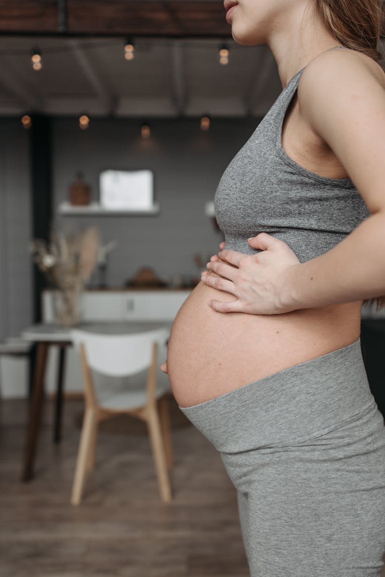 Side view of a pregnant woman wearing grey activewear, standing indoors in a cozy setting.
