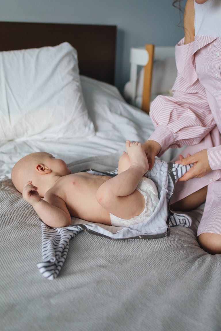Side view of adorable little baby in diaper lying on comfortable bed in light bedroom while crop unrecognizable mother dressing newborn in striped suit at home